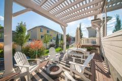 Backyard of a house with a pergola covered patio beside the small door deck. There are wooden armchairs with fire pit in the middle and a dining table with umbrella with a view of a garden and houses.